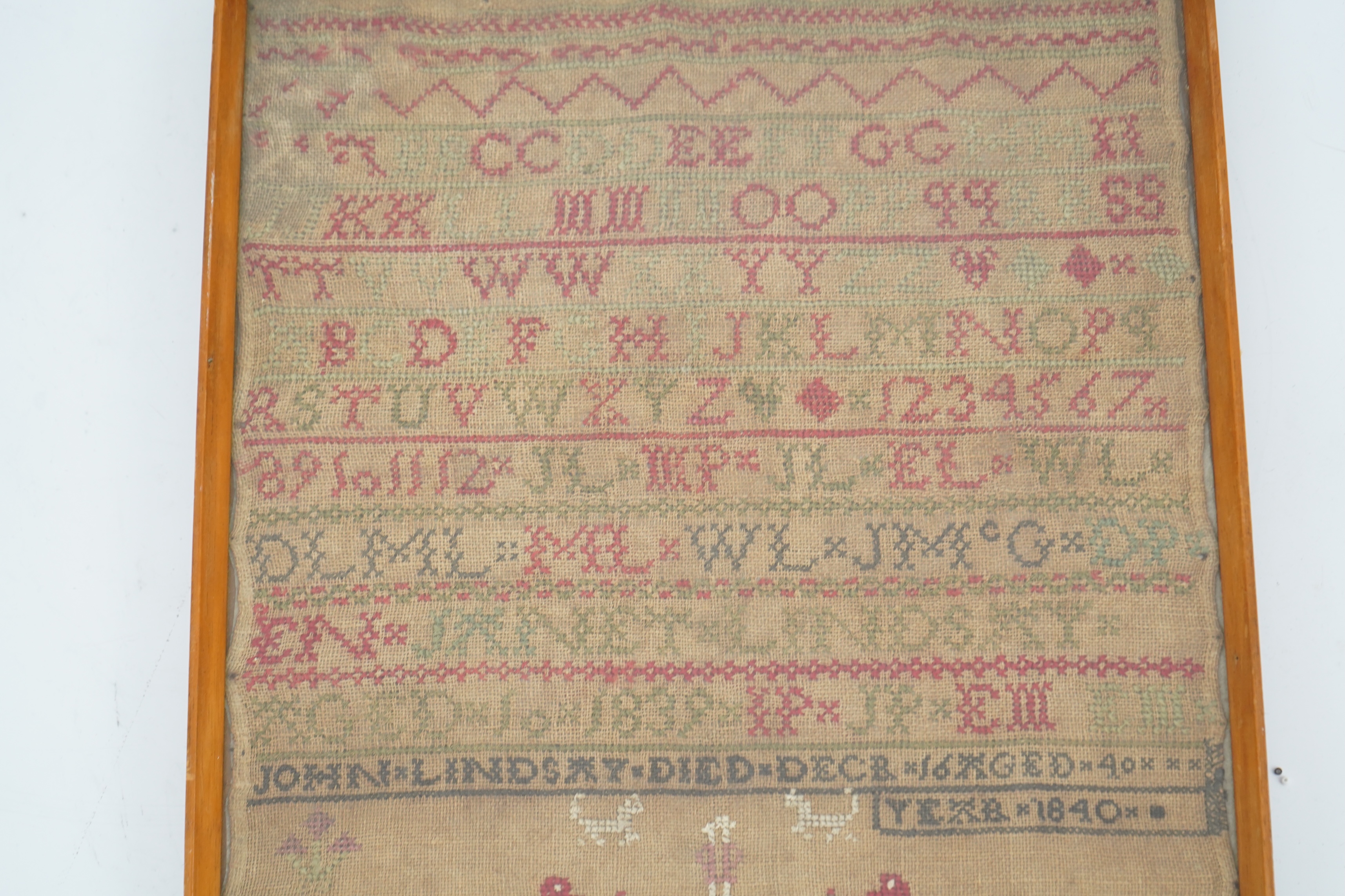 An alphabet cross stitch sampler with a figurative lower spot motif panel and commemorative dedication to John Lindsay, died Dec 16 1840, 27cm wide x 43.5 cm high. Condition - some light staining to linen, the blue and g